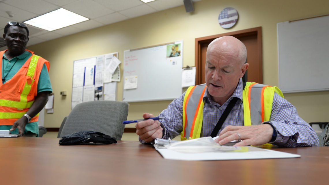 EHS auditor sitting at a table conducting internal audit in a reflective vest with employee standing nearby.