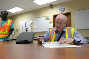 EHS auditor sitting at a table conducting internal audit in a reflective vest with employee standing nearby.